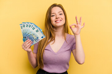 Young caucasian woman holding banknotes isolated on yellow background cheerful and confident...