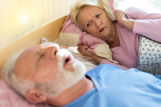 Senior Man Snoring In Bed While Woman Holding Pillow On Ears