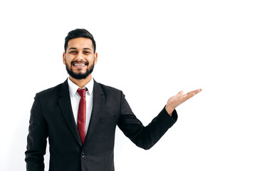 Copy-space for your presentation. Positive elegant arabian or indian man in formal suit, standing over isolated white background, looking at camera, pointing hand to empty space, smiles happily
