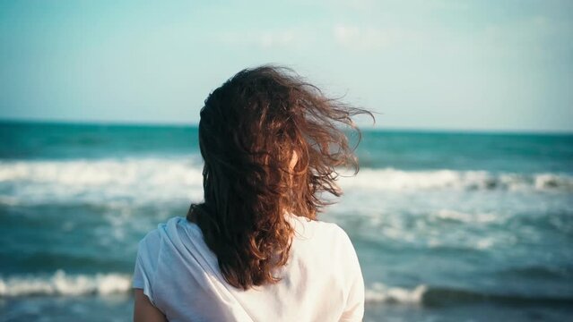 Cinematic back shot of a young woman enjoying the fresh breeze while walking on the beach at sunset time.
