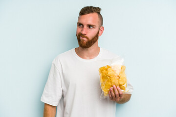 Young caucasian man holding crisps isolated on blue background confused, feels doubtful and unsure.