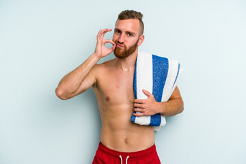 Young caucasian man going to the beach holding a towel isolated on blue background with fingers on lips keeping a secret.