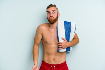 Young caucasian man going to the beach holding a towel isolated on blue background shrugs shoulders and open eyes confused.