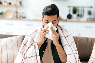 Unhealthy indian or arabian guy sits at home on sofa in living room under plaid, holds napkin near nose, sneezes, suffers from runny nose, feels unwell, needs treatment and doctor consultation - obrazy, fototapety, plakaty