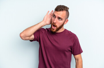Young caucasian man isolated on blue background trying to listening a gossip.
