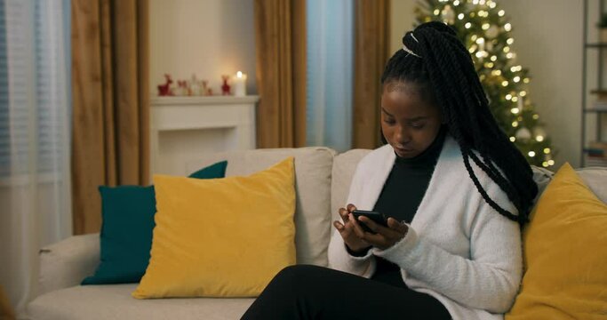 Winter evening. A beautiful girl holds phone in hands and chooses photo for family album. Girl is dressed in black clothes and white cardigan. There are yellow pillows on sofa.