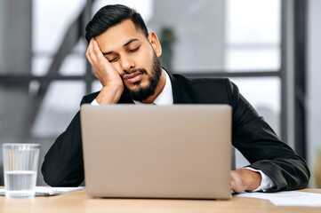 Sleepy arabian or indian business man in a suit, sits at workplace in the office with a laptop, put...