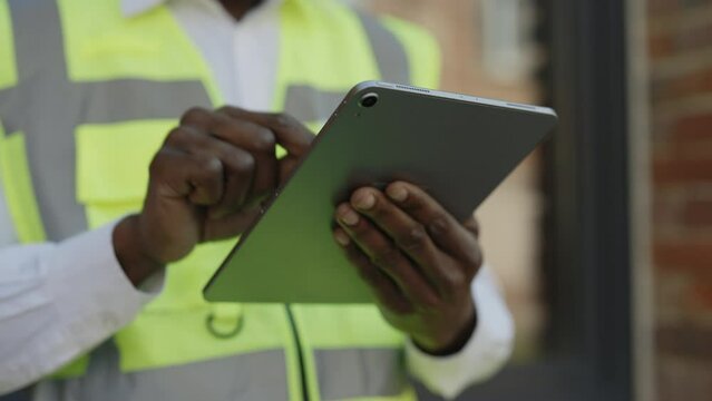 Close up of architect hands using digital tablet near construction site. Unrecognizable african american builder checking drawing online with background of brick wall.