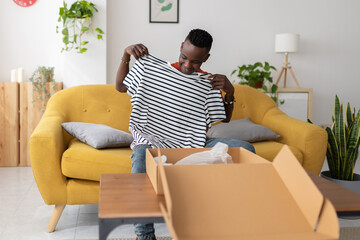 Happy african man unboxing package after delivery - Young adult male holding striped shirt sitting...