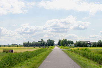 Fototapeta na wymiar Typical Dutch landscape with flat and low land under blue sky in summer, Small countryside road through the polder and water land with green meadow, Small village in Noord Holland, Netherlands.