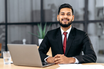 Portrait of a positive young male confident arabian or indian businessman, successful entrepreneur in a suit, sit at a work desk with laptop in modern creative office, looks at camera, smile friendly