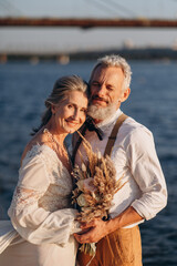 Senior newlyweds hug. Stylish couple of elderly newlyweds stand embracing on river bank.