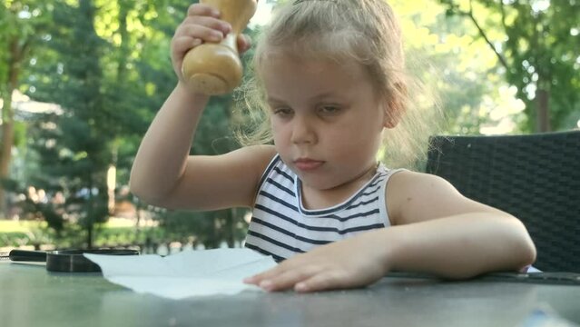 Little girl pours salt from salt shaker onto napkin. Close-up of blonde girl playing with salt shaker in street cafe on the park. Slow motion.