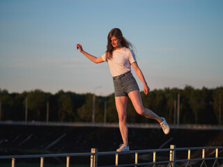Fototapeta na wymiar A young beautiful woman in a white blouse and shorts walks along the railing against the sky, keeping her balance. Happy Summer time