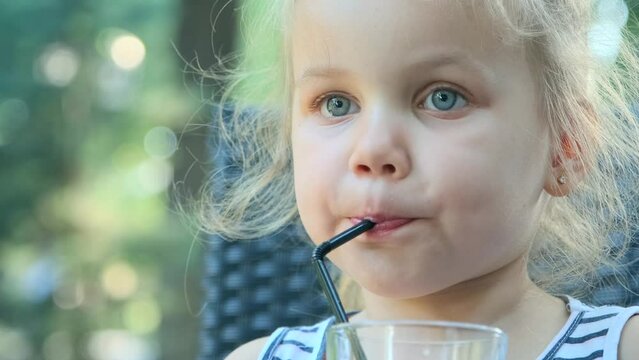 Little girl drinks juice through straw. Close-up portrait of blonde girl drinks juice from glass through cocktail straw sitting in street cafe on the park. Slow motion 