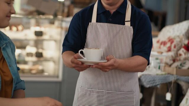 Caucasian male waiter with down syndrome serving a cup of coffee in the cafe to the client at the table. Shot with RED helium camera in 8K. 