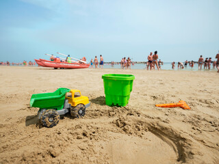 Toy sand truck with bucket and spade on an Italian beach on a sunny summer morning with people in the back as they cool off by the sea. Children's toy by the Adriatic sea, Italy.