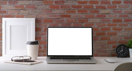 Stylish workplace with laptop computer, coffee cup, stationery and houseplant on white table against brick wall. Blank screen for your advertise text.