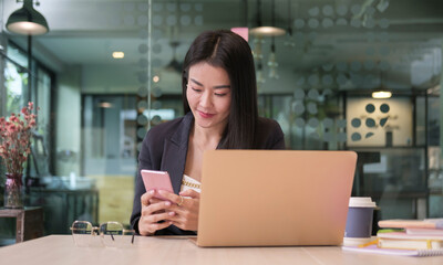 Obraz premium Beautiful young businesswoman using smart phone, reading news, checking social media while sitting in modern office.