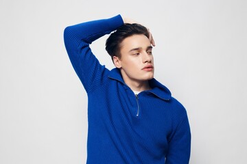 Horizontal studio shot. a handsome stylish man with thick black hair standing on a light gray background in a blue zip-up sweater and looking charmingly into the camera with his hands on his head