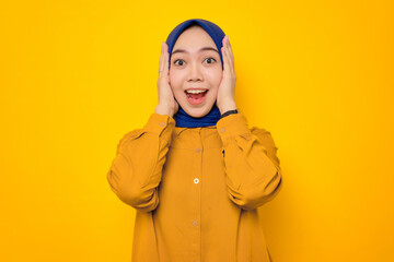 Shocked young Asian Muslim woman dressed in orange shirt looking at camera with open mouth and touching his cheeks isolated on yellow background