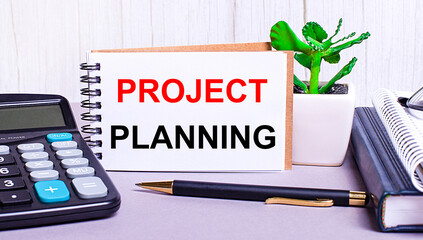 On the desktop are a calculator, diaries, a potted plant, a pen and a notebook with the text PROJECT PLANNING. Business concept. Workplace close up