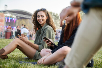 Woman sitting on the grass and using mobile phone at the festival with friends - obrazy, fototapety, plakaty
