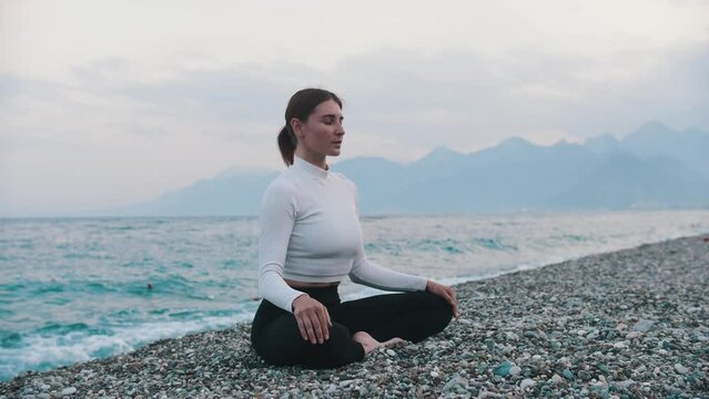 A woman in white sweater sitting in lotus position on the pebble beach by the sea