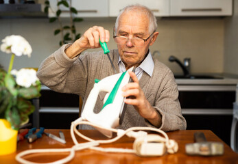 elderly pensioner is reading sitting at table and repairing an iron