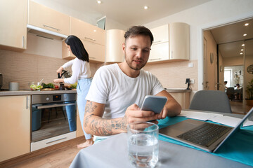 Man at laptop texting on smartphone next to cooking wife