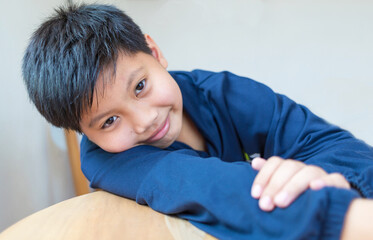 Obraz premium Cute Asian boy with white skin smiling looking in camera happily resting chins on hands-on wooden table. Close-up portrait of a cute child.