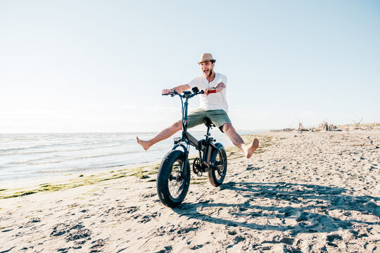 Young man on bicycle having fun with electric bike - carefree boy having fun and smiling on bicycle on the beach on a sunny day - freedom concept