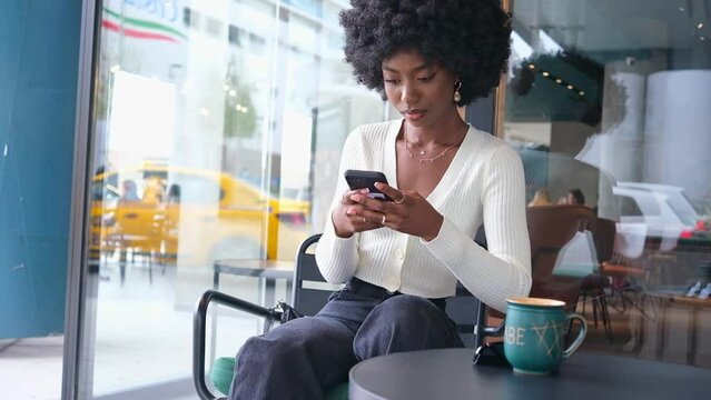 Happy African American Woman Sitting In Cafe With Coffee Using Smart Phone Typing Scrolling Outside