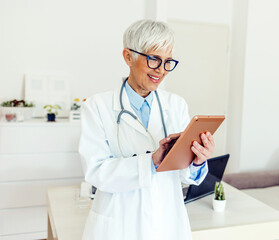 Smiling mature female doctor with stethoscope using tablet at medical clinic. Happy healthcare worker using computer at modern hospital.