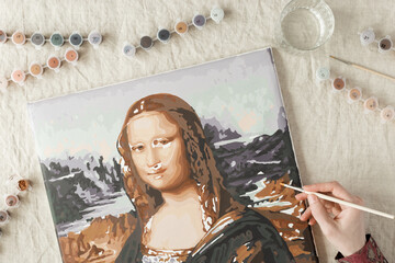 Painting by numbers with acrylic paints. Woman coloring Mona Lisa picture by numbers, hand with brush and paints of different colors. Creative hobby for relaxation, leisure activity at home