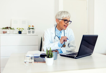 A mature women doctor sitting at the workplace and looking at the laptop during the day.  Portrait of a female doctor wearing white uniform using laptop to talk to patient online.