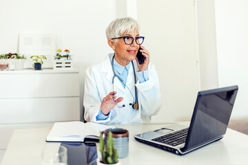 Confident mature doctor in white lab coat talking on the phone and smiling while sitting in her office. Beautiful mature female physician sitting at the table with laptop and having phone conversation