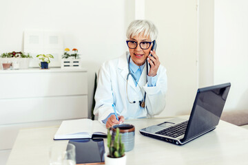 Doctor is on the phone and checking medical records. Female senior doctor sitting behind desk in office, using smart phone to talk with patients.