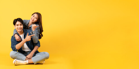 Two Young good looking asian couple isolated on yellow background sitting and relax pointing at...