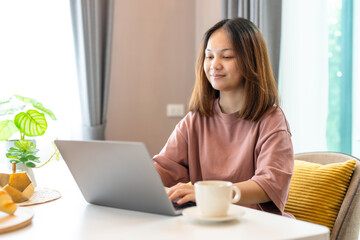 young Asian woman with long hair sitting in the living room on the sofa using a laptop to video call a coworker. During the conversation, typed a message as well. Work from home.