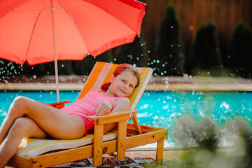 Pretty teen girl with wet hair and pink summer swimsuit posing on beach chair with red umbrella...