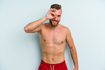 Young caucasian man wearing a swimsuit isolated on blue background excited keeping ok gesture on eye.