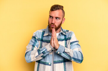 Young caucasian man isolated on yellow background praying, showing devotion, religious person looking for divine inspiration.