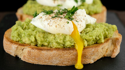Avocado toast with poached egg close up. Cutting poached egg with runny egg yolk over bread toast with mashed avocado spread