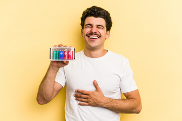 Young caucasian man holding batteries isolated on yellow background laughs out loudly keeping hand on chest.