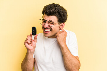 Young caucasian man holding car keys isolated on yellow background covering ears with hands.