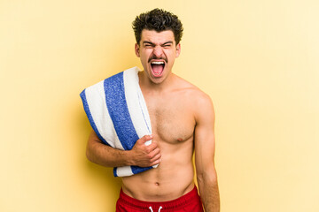 Young caucasian man holding beach towel isolated on yellow background screaming very angry and aggressive.