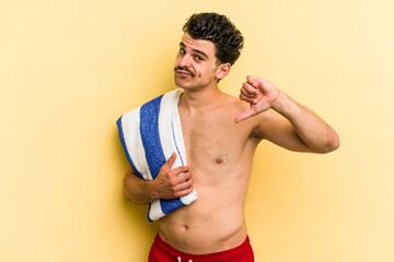 Young caucasian man holding beach towel isolated on yellow background showing a dislike gesture, thumbs down. Disagreement concept.