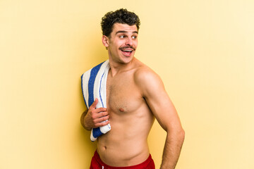 Young caucasian man holding beach towel isolated on yellow background looks aside smiling, cheerful and pleasant.