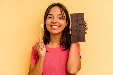 Young hispanic woman washing teeth after eating chocolate isolated on yellow background